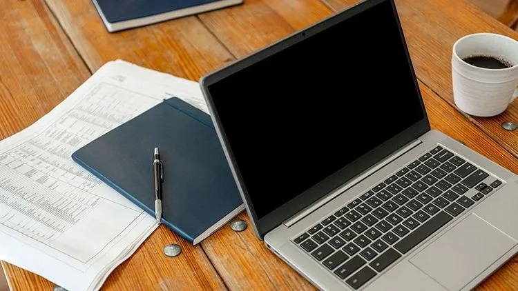 Geöffneter Laptop liegt neben Unterlagen, Notizbuch und Kaffeetasse auf einem Holztisch.