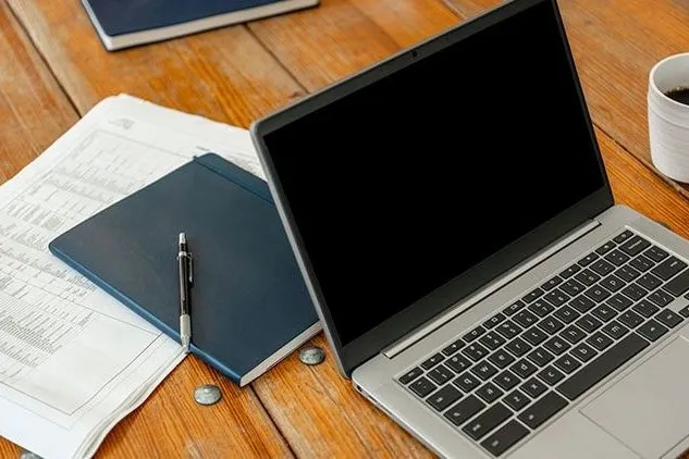 Geöffneter Laptop liegt neben Unterlagen, Notizbuch und Kaffeetasse auf einem Holztisch.