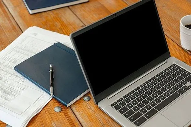 Geöffneter Laptop liegt neben Unterlagen, Notizbuch und Kaffeetasse auf einem Holztisch.