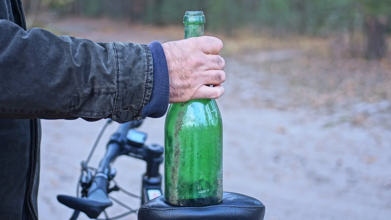 Person hält eine grüne Flasche neben einem Fahrrad auf einem Waldweg.