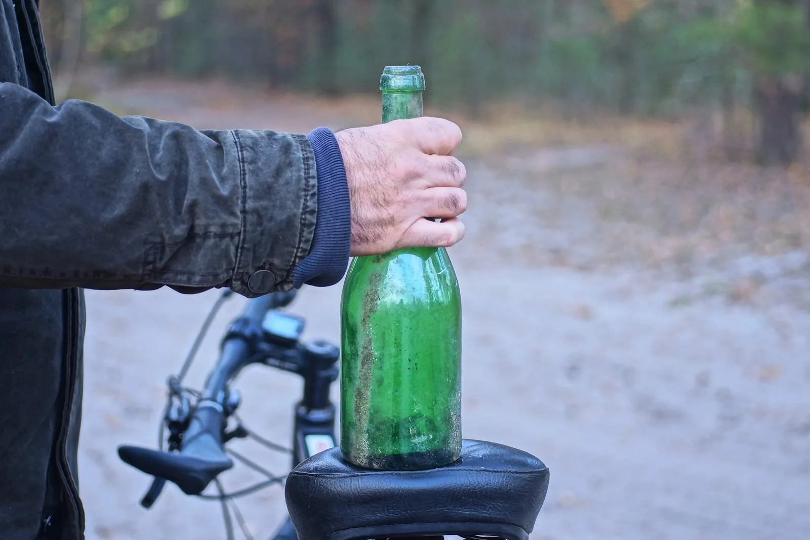 Person hält eine grüne Flasche neben einem Fahrrad auf einem Waldweg.