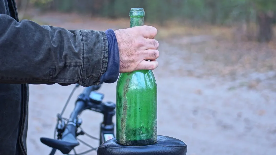 Person hält eine grüne Flasche neben einem Fahrrad auf einem Waldweg.