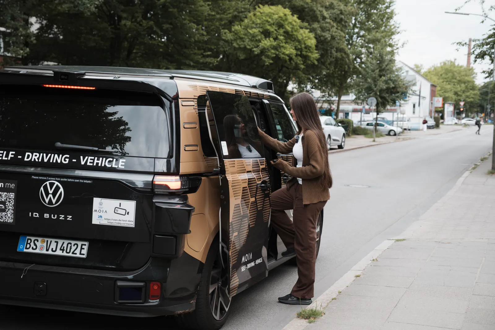 Autonomer VW ID. Buzz von Moia steht am Straßenrand, während eine Frau an der geöffneten Schiebetür einsteigt.