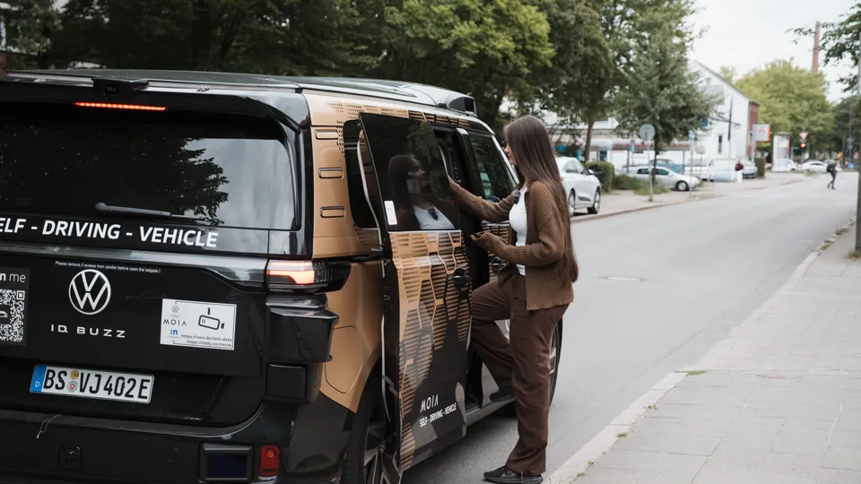 Autonomer VW ID. Buzz von Moia steht am Straßenrand, während eine Frau an der geöffneten Schiebetür einsteigt.