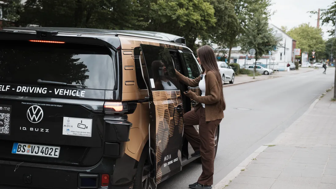 Autonomer VW ID. Buzz von Moia steht am Straßenrand, während eine Frau an der geöffneten Schiebetür einsteigt.