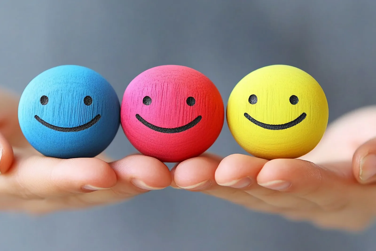 Zwei Hände die drei Smileys halten: ein blauer, ein roter und ein gelber. Die Smileys lächeln.