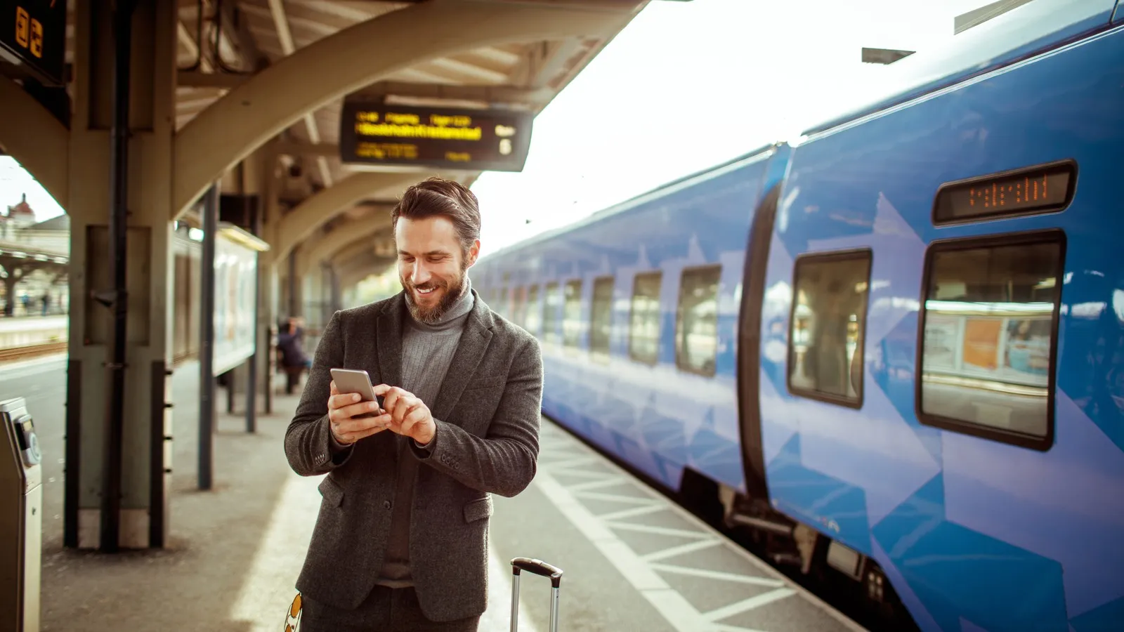 Geschäftsreisender steht mit Smartphone und Koffer am Bahnsteig neben einem Zug.