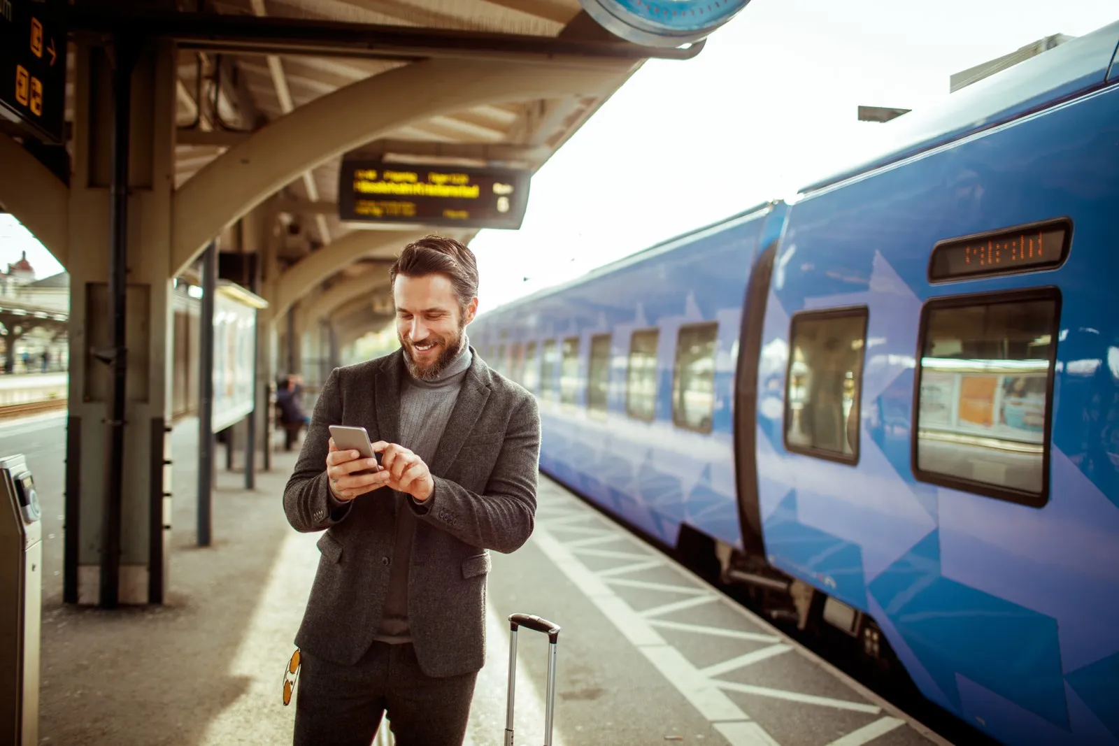 Geschäftsreisender steht mit Smartphone und Koffer am Bahnsteig neben einem Zug.