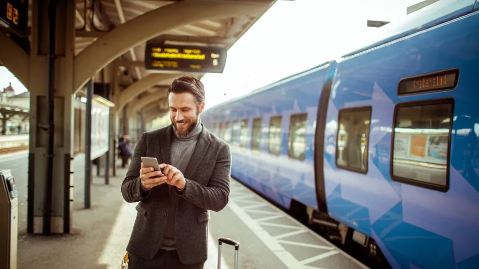 Geschäftsreisender steht mit Smartphone und Koffer am Bahnsteig neben einem Zug.