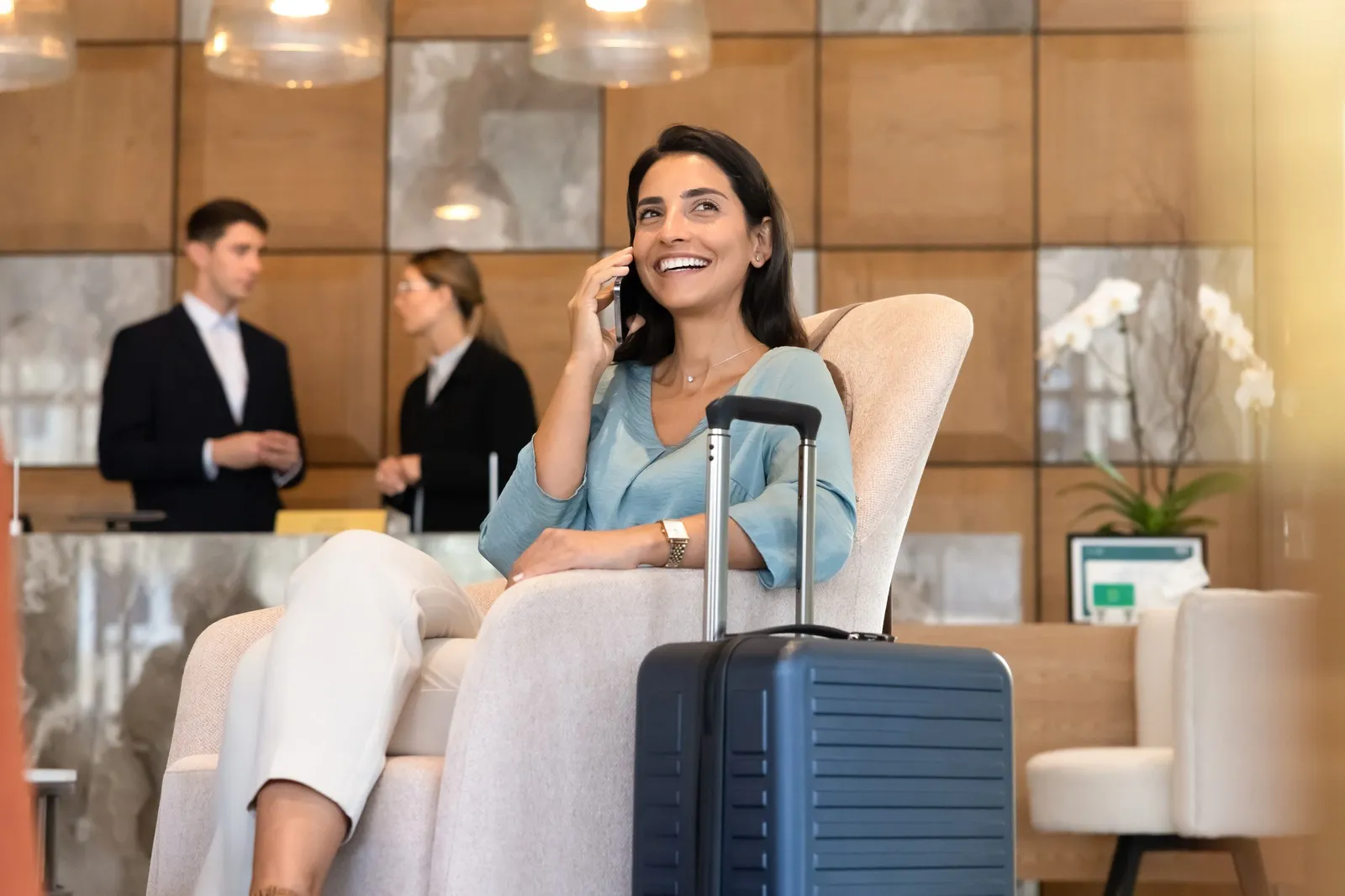 Eine Geschäftsfrau sitzt mit Koffer in einer Hotellobby und telefoniert, im Hintergrund stehen zwei Personen an der Rezeption.