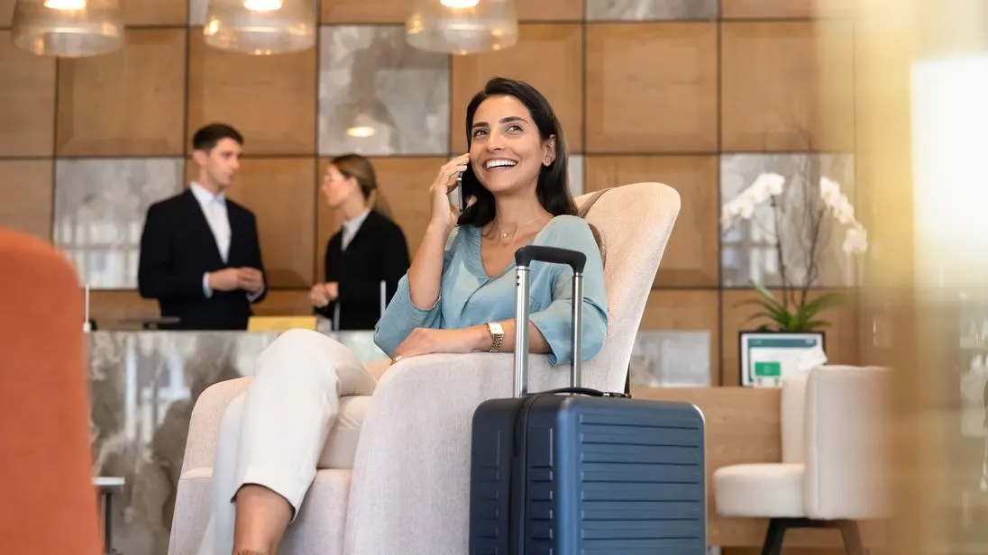 Eine Geschäftsfrau sitzt mit Koffer in einer Hotellobby und telefoniert, im Hintergrund stehen zwei Personen an der Rezeption.