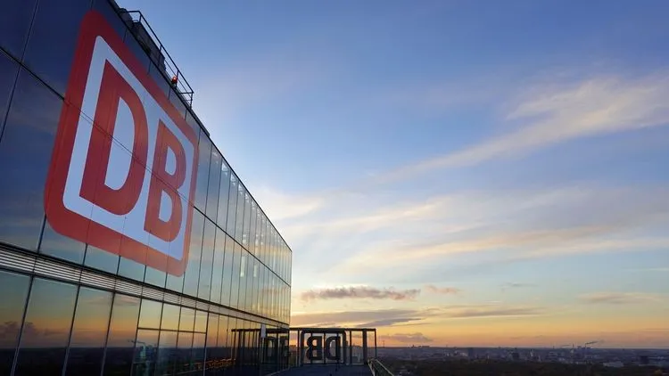 DB-Logo an der Glasfassade eines Bürogebäudes bei Sonnenuntergang.