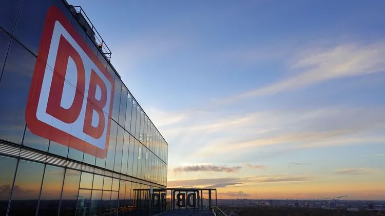 DB-Logo an der Glasfassade eines Bürogebäudes bei Sonnenuntergang.