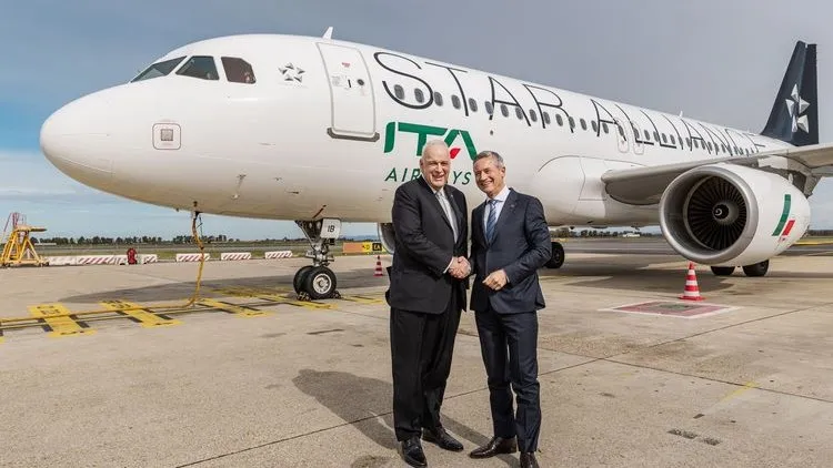Zwei Männer in Anzügen stehen auf dem Rollfeld vor einem Flugzeug von ITA Airways und geben sich die Hand. Das Passagierflugzeug trägt die Lackierung der Star Alliance.