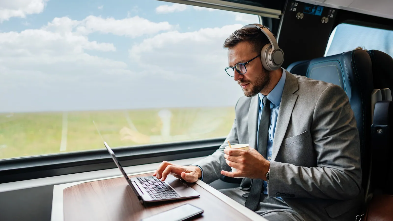Geschäftsreisender sitzt im Zug am Tisch, arbeitet am Laptop und trägt Kopfhörer.