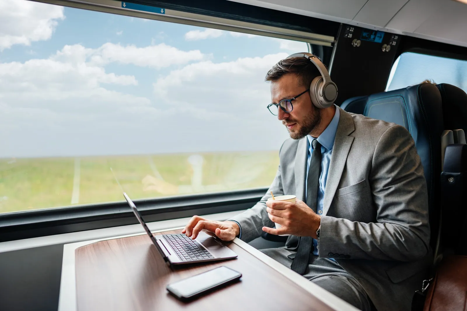 Geschäftsreisender sitzt im Zug am Tisch, arbeitet am Laptop und trägt Kopfhörer.