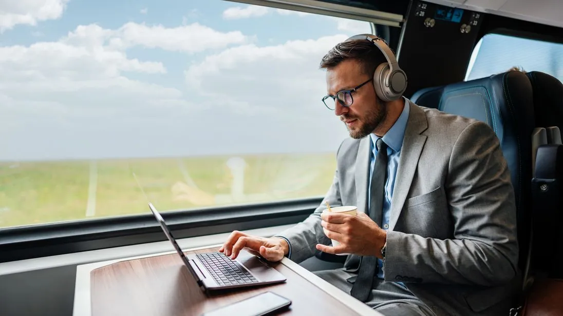 Geschäftsreisender sitzt im Zug am Tisch, arbeitet am Laptop und trägt Kopfhörer.