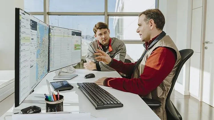 Zwei Mitarbeitende in Arbeitskleidung sitzen an einem Schreibtisch und schauen gemeinsam auf Monitore mit digitalen Flotten- und Fahrzeugdaten.