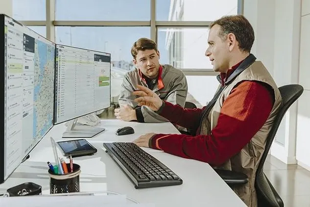 Zwei Mitarbeitende in Arbeitskleidung sitzen an einem Schreibtisch und schauen gemeinsam auf Monitore mit digitalen Flotten- und Fahrzeugdaten.