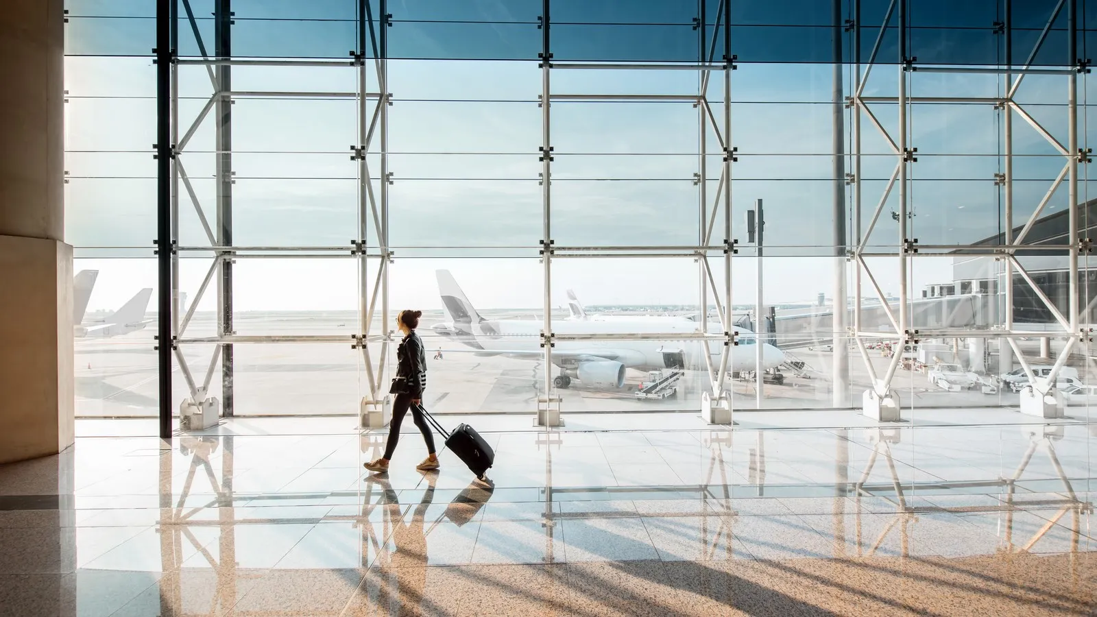 Eine Person mit Rollkoffer geht durch ein helles Flughafenterminal. Durch die große Glasfront sind Flugzeuge auf dem Vorfeld zu sehen.