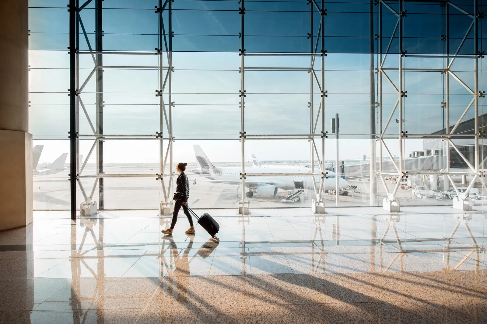 Eine Person mit Rollkoffer geht durch ein helles Flughafenterminal. Durch die große Glasfront sind Flugzeuge auf dem Vorfeld zu sehen.