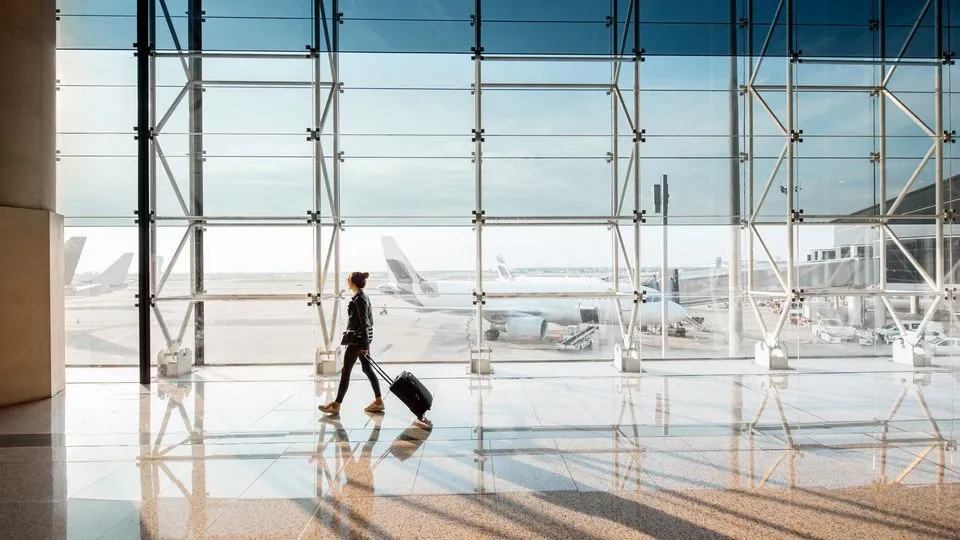 Eine Person mit Rollkoffer geht durch ein helles Flughafenterminal. Durch die große Glasfront sind Flugzeuge auf dem Vorfeld zu sehen.