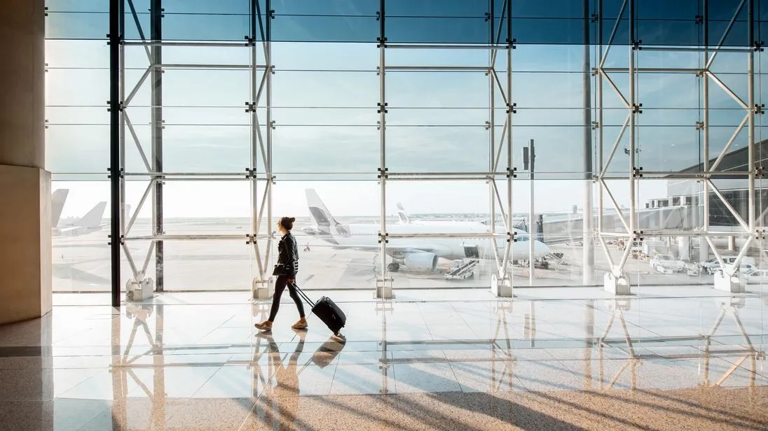 Eine Person mit Rollkoffer geht durch ein helles Flughafenterminal. Durch die große Glasfront sind Flugzeuge auf dem Vorfeld zu sehen.