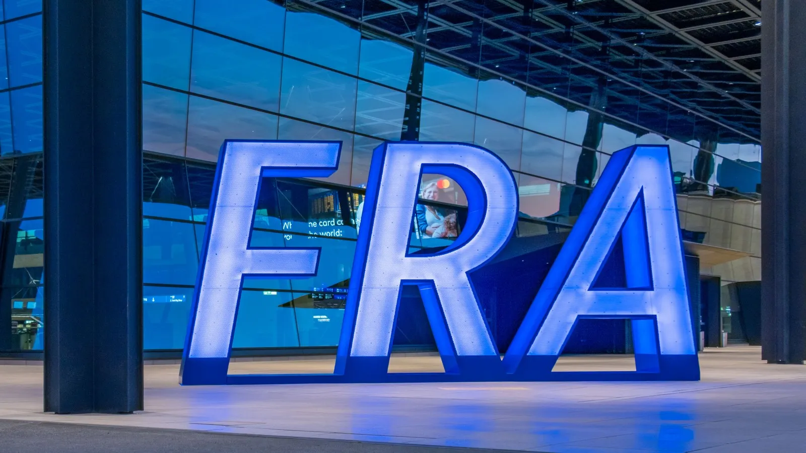 Vor dem Terminal des Frankfurter Flughafens steht ein großes beleuchtetes „FRA“-Logo. Im Hintergrund ist die Glasfassade des Flughafengebäudes zu sehen.