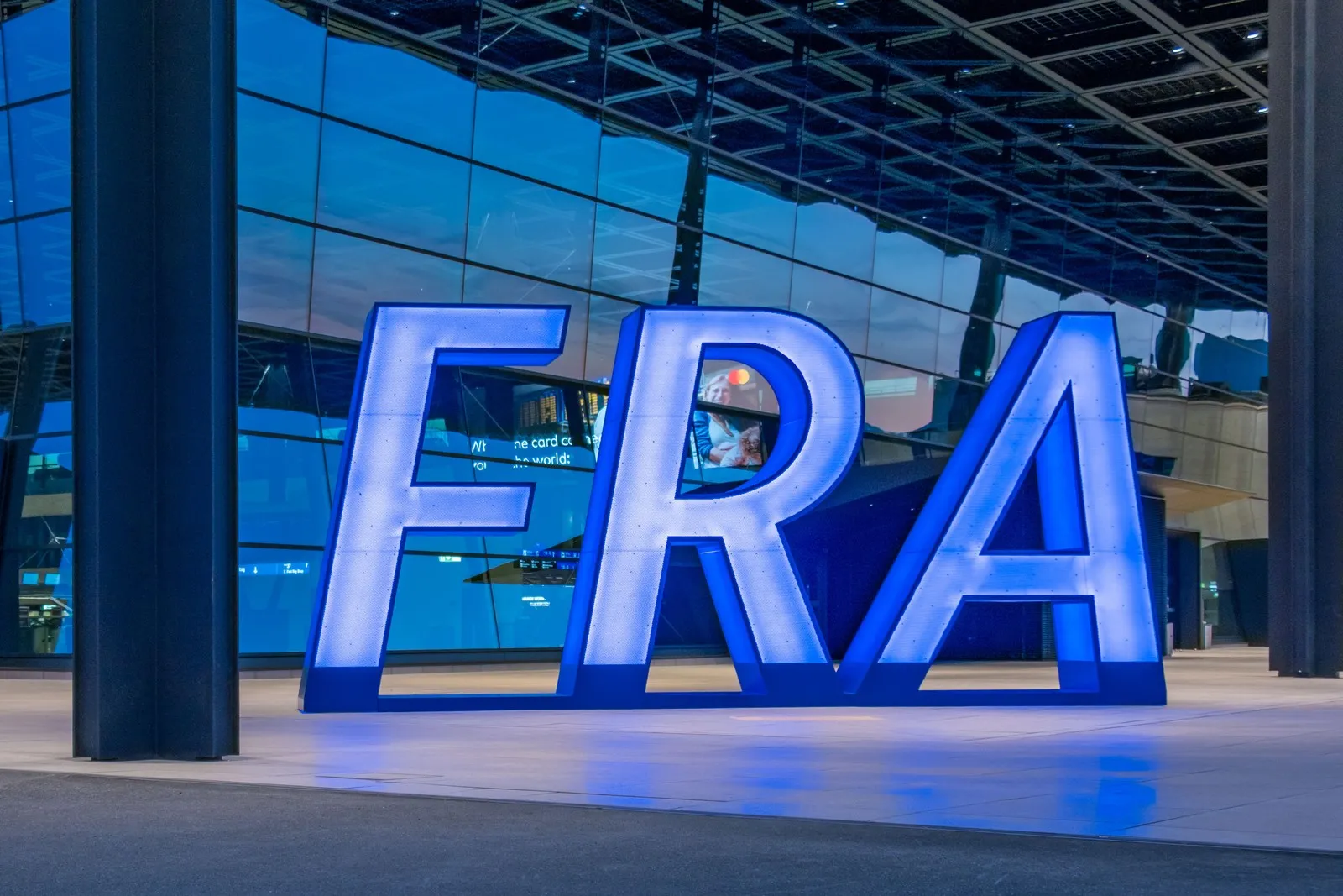 Vor dem Terminal des Frankfurter Flughafens steht ein großes beleuchtetes „FRA“-Logo. Im Hintergrund ist die Glasfassade des Flughafengebäudes zu sehen.