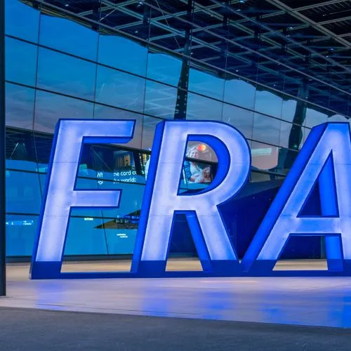 Vor dem Terminal des Frankfurter Flughafens steht ein großes beleuchtetes „FRA“-Logo. Im Hintergrund ist die Glasfassade des Flughafengebäudes zu sehen.