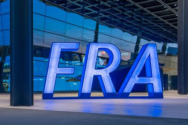 Vor dem Terminal des Frankfurter Flughafens steht ein großes beleuchtetes „FRA“-Logo. Im Hintergrund ist die Glasfassade des Flughafengebäudes zu sehen.