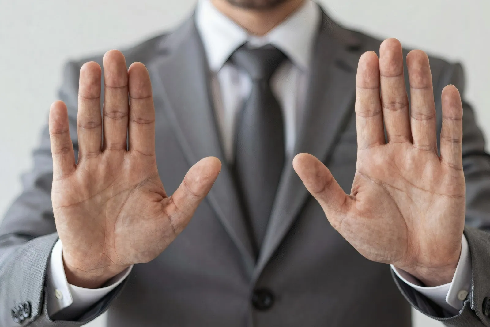 Businessmann in einem grauen Anzug mit Krawatte gestikuliert mit seinen Händen eine ablehnende Haltung, Hintergrund ist eine weiße Wand.