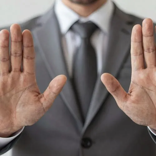 Businessmann in einem grauen Anzug mit Krawatte gestikuliert mit seinen Händen eine ablehnende Haltung, Hintergrund ist eine weiße Wand.