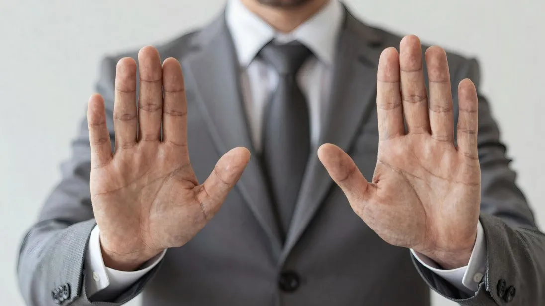 Businessmann in einem grauen Anzug mit Krawatte gestikuliert mit seinen Händen eine ablehnende Haltung, Hintergrund ist eine weiße Wand.