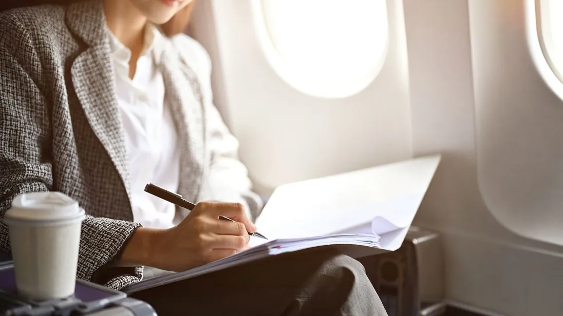Eine Frau sitzt im Flugzeug in einem Meeting und macht sich in ihrer Mappe mit einem Stift Notizen.