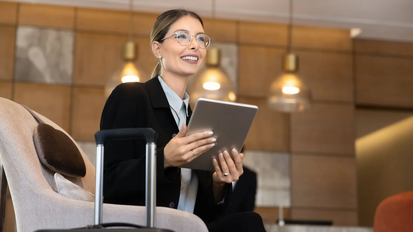 Eine Frau sitzt mit ihrem Koffer in der Hotellobby, nutzt ein Tablet, um während einer Geschäftsreise berufliche Aufgaben zu erledigen.