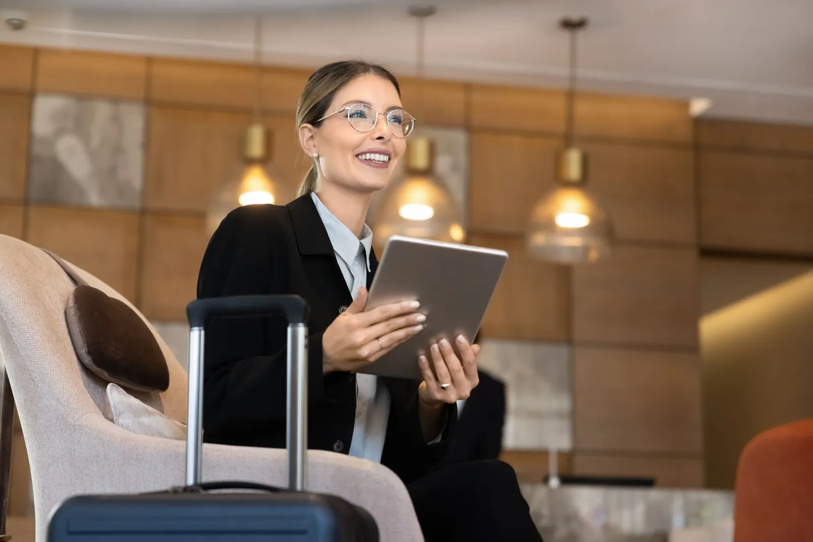 Eine Frau sitzt mit ihrem Koffer in der Hotellobby, nutzt ein Tablet, um während einer Geschäftsreise berufliche Aufgaben zu erledigen.