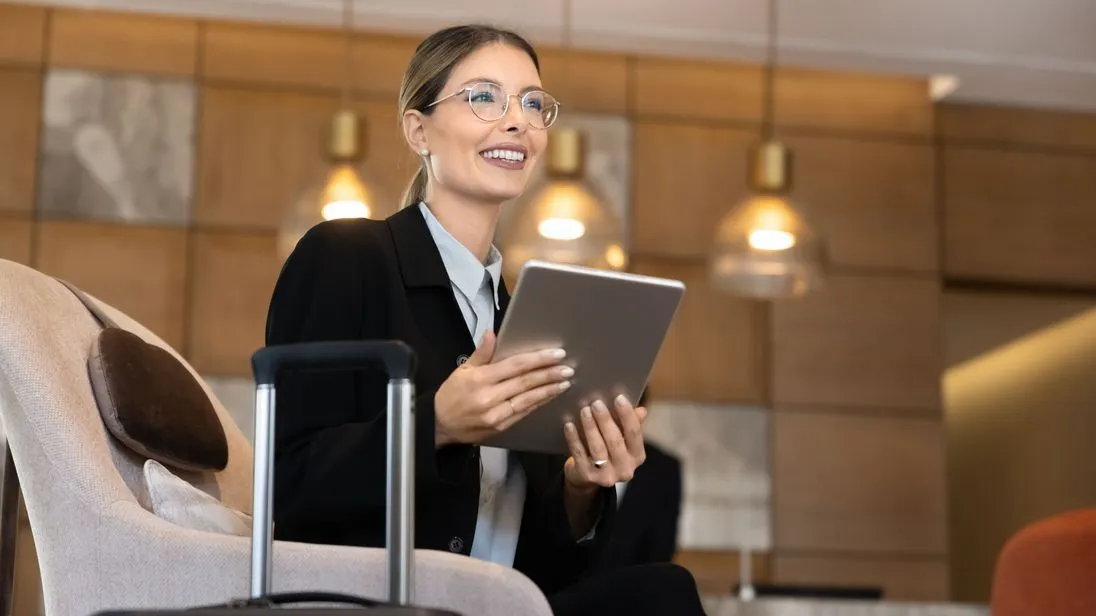 Eine Frau sitzt mit ihrem Koffer in der Hotellobby, nutzt ein Tablet, um während einer Geschäftsreise berufliche Aufgaben zu erledigen.
