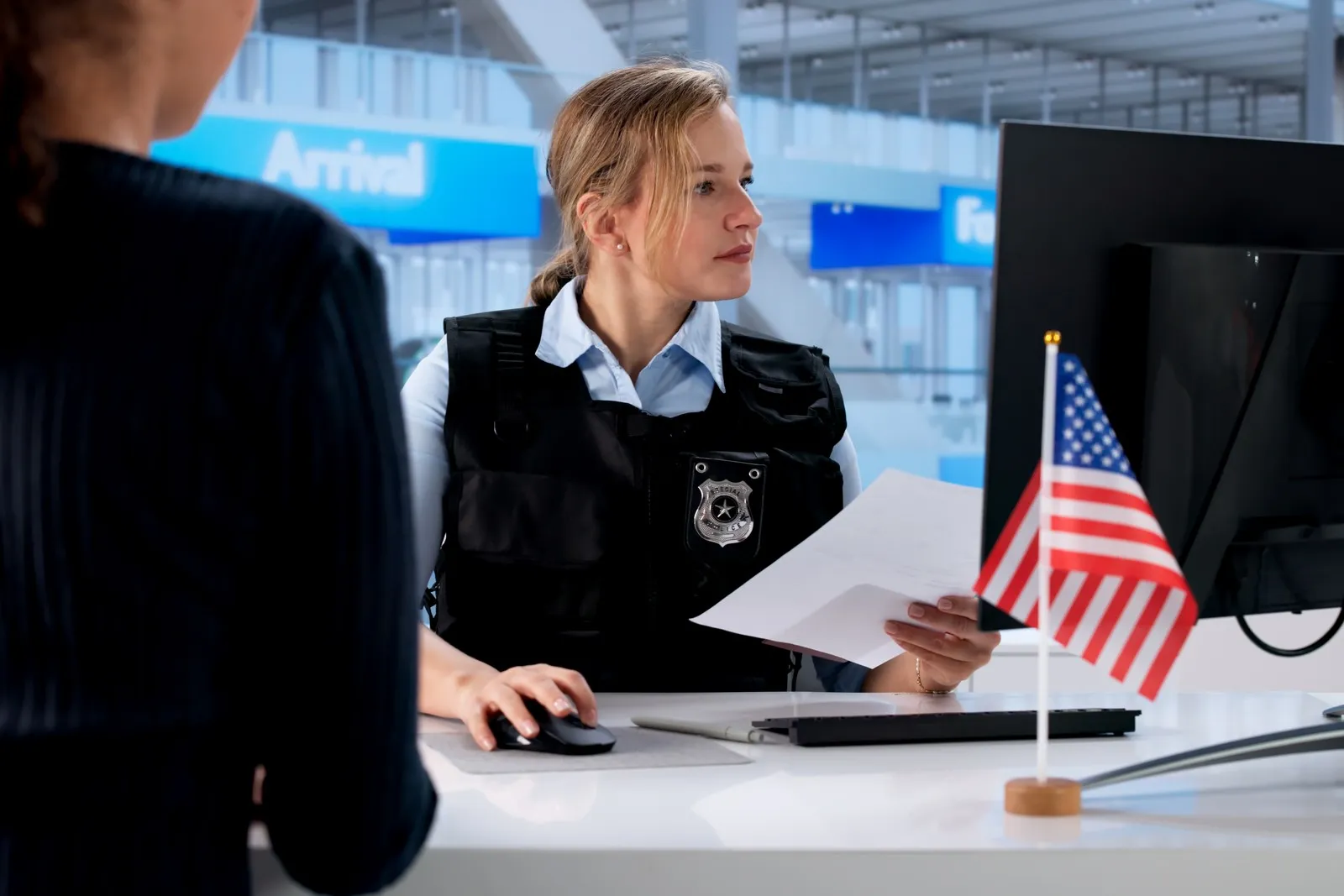 Sicherheitsbeamtin in Uniform bei der Dokumentenkontrolle an einem US-Grenzschalter. Im Vordergrund eine US-Flagge, im Hintergrund Flughafen-Terminal. Symbolbild für die verschärften Sicherheitsmaßnahmen und verstärkten Kontrollen bei der Einreise in die USA 2026.