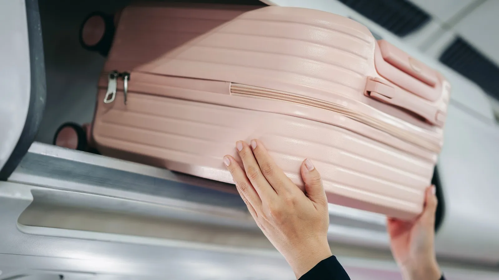 Hände schieben einen pinken Handgepäckskoffer in die Gepäckablage im Flugzeug.