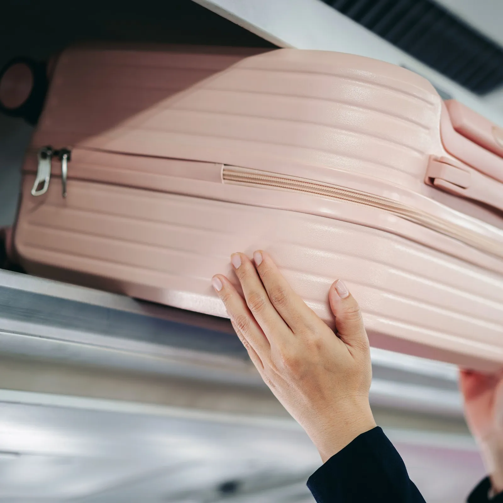 Hände schieben einen pinken Handgepäckskoffer in die Gepäckablage im Flugzeug.