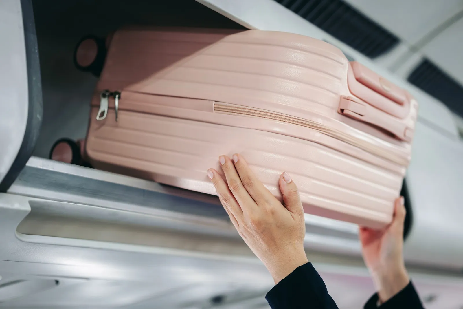 Hände schieben einen pinken Handgepäckskoffer in die Gepäckablage im Flugzeug.