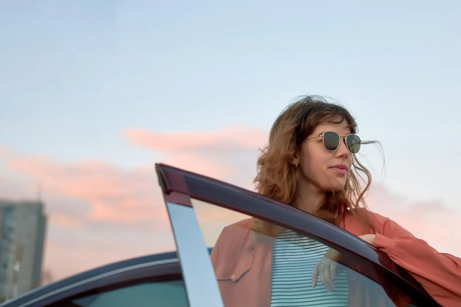 Eine Frau mit Sonnenbrille steht an einer geöffneten Autotür und blickt zur Seite. Im Hintergrund ist ein unscharfer Himmel mit rosa Wolken zu sehen.