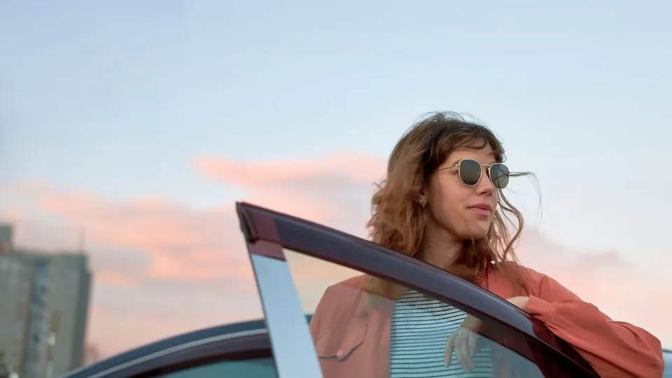 Eine Frau mit Sonnenbrille steht an einer geöffneten Autotür und blickt zur Seite. Im Hintergrund ist ein unscharfer Himmel mit rosa Wolken zu sehen.