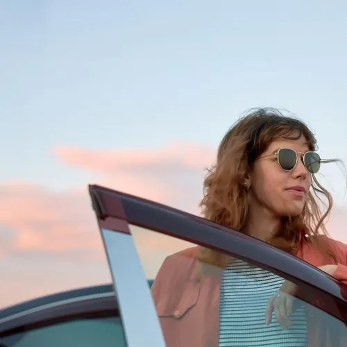 Eine Frau mit Sonnenbrille steht an einer geöffneten Autotür und blickt zur Seite. Im Hintergrund ist ein unscharfer Himmel mit rosa Wolken zu sehen.