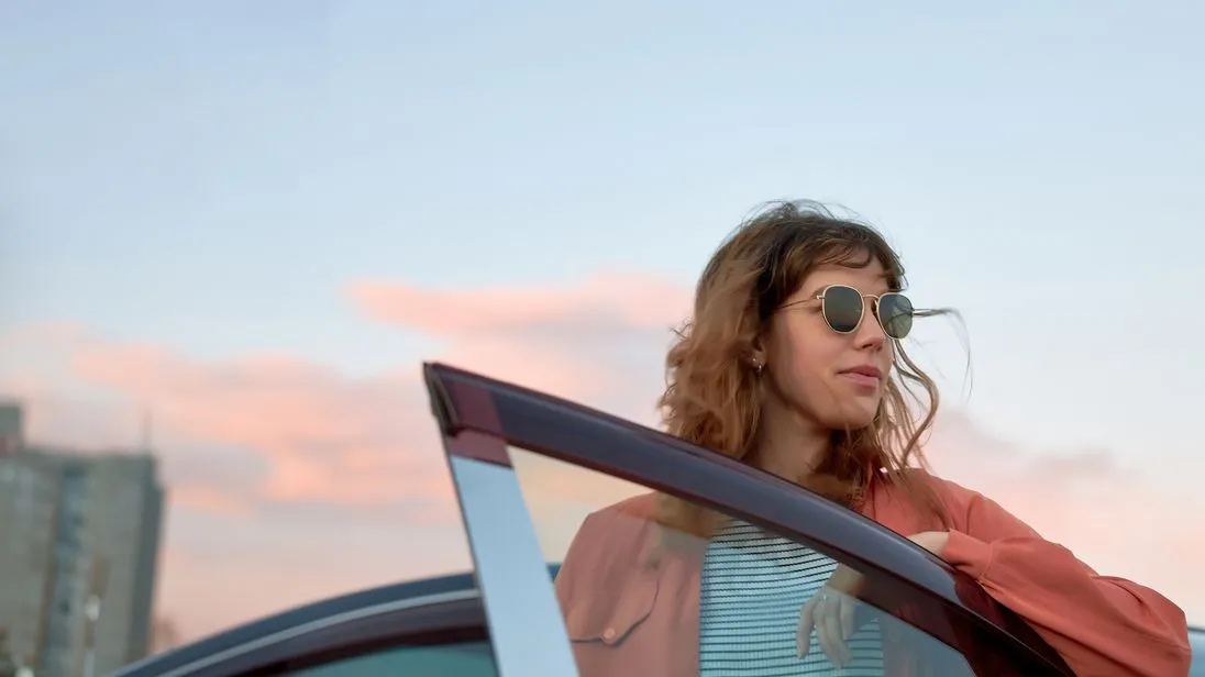 Eine Frau mit Sonnenbrille steht an einer geöffneten Autotür und blickt zur Seite. Im Hintergrund ist ein unscharfer Himmel mit rosa Wolken zu sehen.