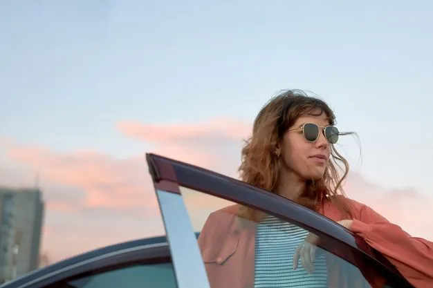Eine Frau mit Sonnenbrille steht an einer geöffneten Autotür und blickt zur Seite. Im Hintergrund ist ein unscharfer Himmel mit rosa Wolken zu sehen.