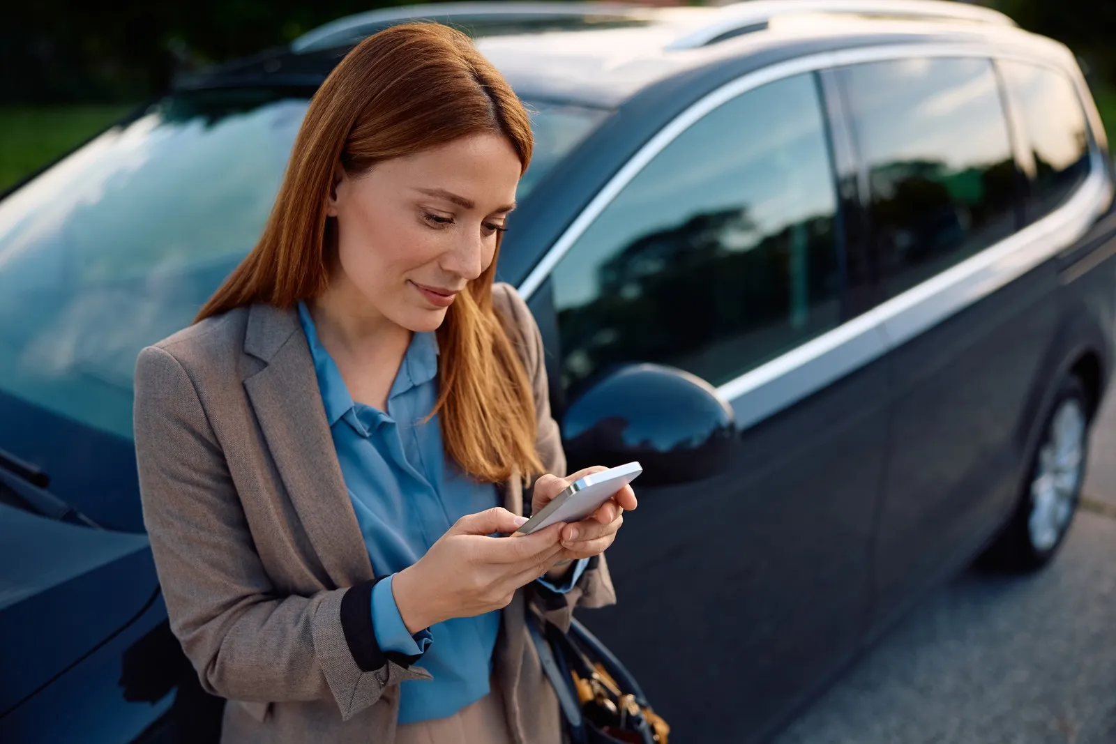 Eine rothaarige Frau lehnt an ihrem Auto oder Mietwagen und schaut auf ihr Handy.