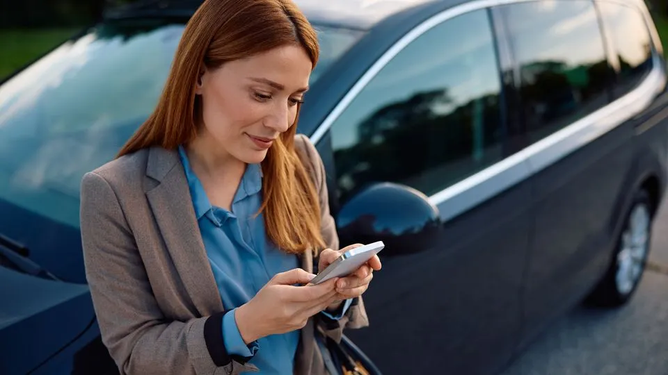 Eine rothaarige Frau lehnt an ihrem Auto oder Mietwagen und schaut auf ihr Handy.