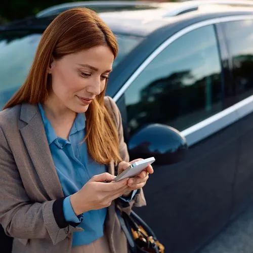 Eine rothaarige Frau lehnt an ihrem Auto oder Mietwagen und schaut auf ihr Handy.